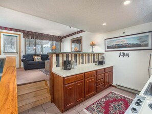 A compact kitchen with a central island, a dining area with a couch, and a framed landscape painting on the wall. - Sunrise A-118:Ski-In/Ski-Out Sunday River Condo (Newry)