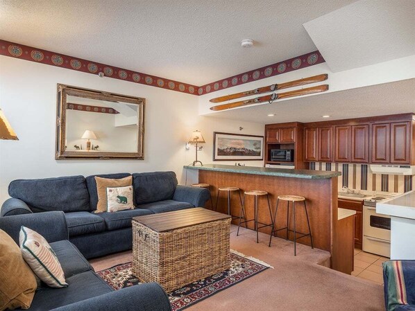 En stue med sofa, sofabord og et køkkenområde med bar samt barstole.