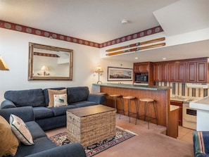 A living room with a sofa, a coffee table, and a kitchen area with a bar and stools. - Sunrise A-118:Ski-In/Ski-Out Sunday River Condo (Newry)