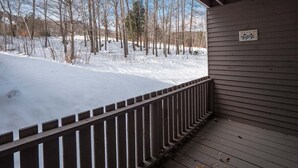 A wooden deck with a railing, a snow-covered ground, and leafless trees in the background.