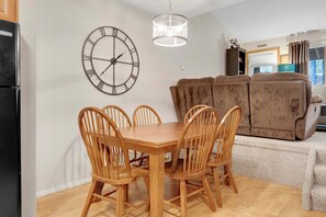 A dining area with a wooden table and chairs, a wall clock, a hanging light fixture, and a brown sofa in the background.