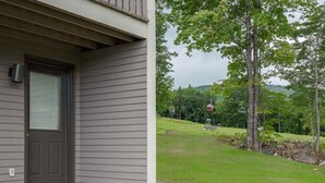 A house with a dark front door, a small porch, and a grassy yard with trees. - Ski-In/Ski-Out Sunday River Condo: Sunrise A-133 (Newry)