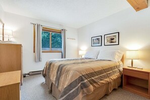 Une chambre à coucher avec un lit, une table de chevet, une lampe, une fenêtre avec des rideaux et deux tableaux encadrés accrochés au mur.