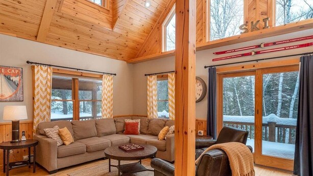 Un intérieur de chalet chaleureux avec un plafond en bois, de grandes fenêtres, un canapé d’angle, une table basse et une cheminée.