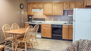 A compact kitchen with wooden cabinets, a stove, a dishwasher, and a refrigerator. There is a dining table with chairs and a clock on the wall.