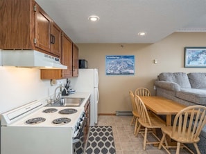 A compact kitchen with wooden cabinets, a stove, and a dining area with a table and chairs. - Ski-In/Ski-Out Sunday River Condo: Sunrise A-101 (Newry)