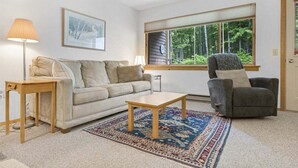Una sala de estar con sofá, sillón, mesa de centro, lámpara y un ventanal con vistas a árboles.