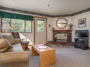 A living room with a sofa, a coffee table, a fireplace, a television, and a door. - Brookside 1 B-104 - Sunday River Slopeside Condominium (Newry)