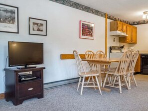Un salon avec un téléviseur, une table à manger en bois et des chaises, un coin cuisine avec des meubles en bois, et des tableaux encadrés accrochés au mur.
