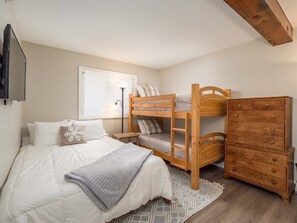 Un dormitorio con una cama nido, un aparador, una lámpara de noche y una ventana con persianas.