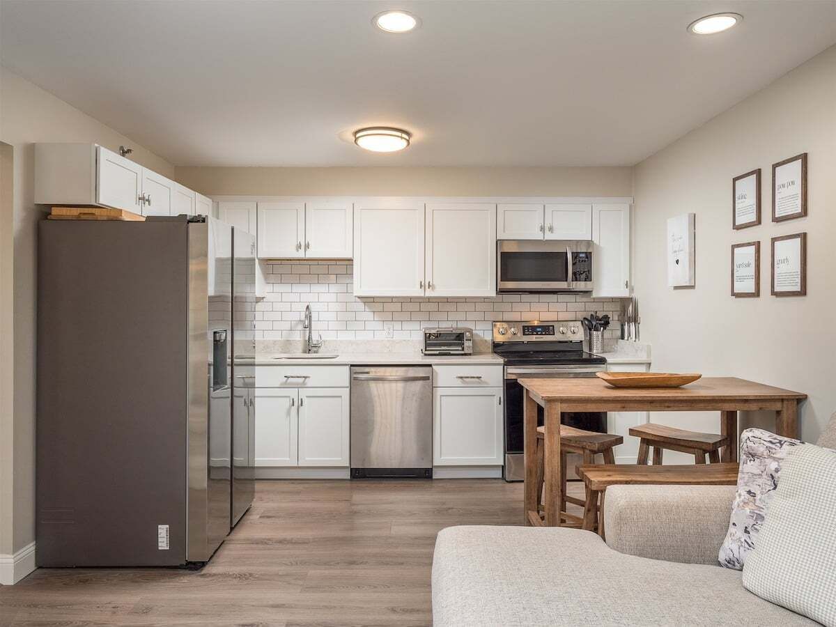 Una cocina compacta con armarios blancos, un refrigerador de acero inoxidable, un microondas y un lavavajillas. Hay una mesa de madera con bancos.