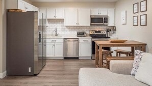 Una cocina compacta con armarios blancos, un refrigerador de acero inoxidable, un microondas y un lavavajillas. Hay una mesa de madera con bancos.