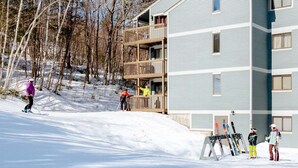 Un edificio de varios pisos con un balcón en el segundo nivel, rodeado de tierra cubierta de nieve y árboles sin hojas.