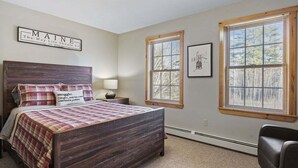 A bedroom with a wooden bed, a nightstand, a lamp, a chair, and two windows with blinds.