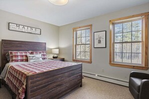 A bedroom with a wooden bed, a nightstand, a lamp, a chair, and two windows with blinds.