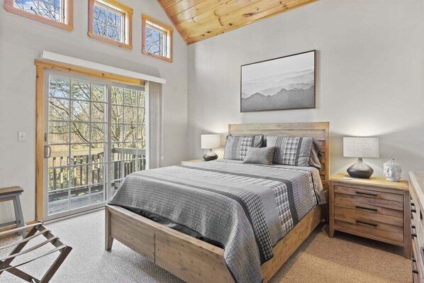 A bedroom with a large window, a bed, a nightstand, and a painting on the wall.