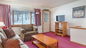 A cozy room with a sofa, armchair, wooden coffee table, TV stand, and a door with a sign. - Ski-In/Ski-Out Sunday River Condo: Sunrise A-120 (Newry)