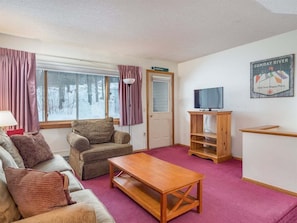 A cozy room with a sofa, armchair, wooden coffee table, TV stand, and a door with a sign. - Ski-In/Ski-Out Sunday River Condo: Sunrise A-120 (Newry)
