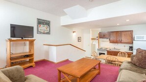 A living room with a TV, wooden entertainment unit, and a dining area with a kitchen in the background. - Ski-In/Ski-Out Sunday River Condo: Sunrise A-120 (Newry)