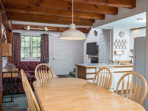 A dining area with a wooden table and chairs, a hanging light fixture, a kitchen with a refrigerator, and a window with curtains. - Riverbend 41: Two bedroom Condo (Bethel)