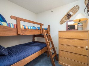 Una cama individual superpuesta con escalera, un aparador de madera y un ventilador fijado a la pared.