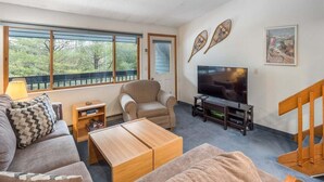 Un salón con sofá, sillón, televisor, mesa de centro de madera y vistas a árboles a través de amplias ventanas.
