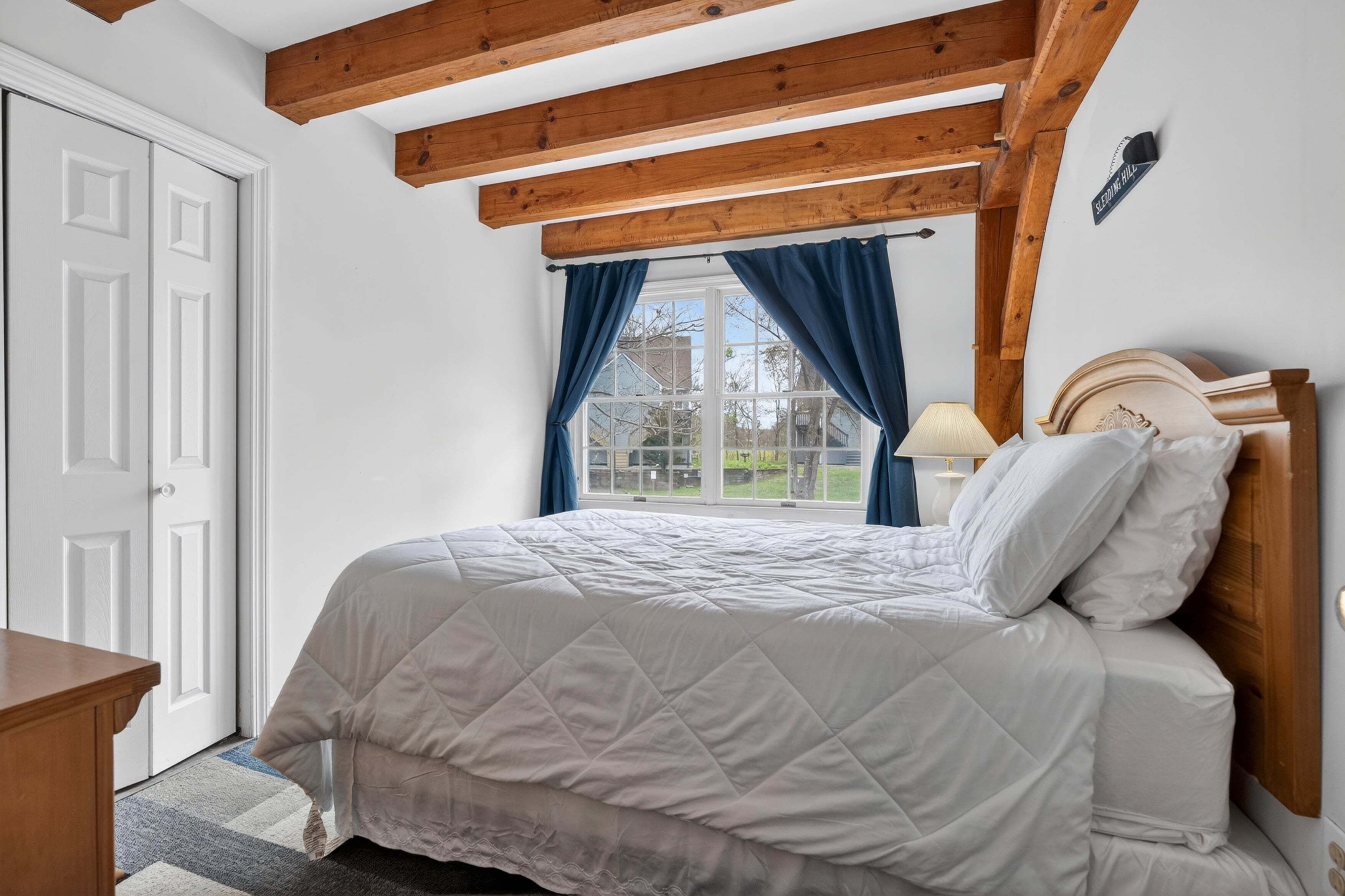 Een slaapkamer met een bed, houten balken in het plafond, een raam met gordijnen, een nachtkastje en een lamp.