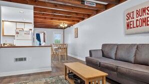 A living room with a brown leather sofa, a wooden coffee table, and a dining area with a table and chairs. - Riverbend 29 - Post and Beam Condo (Bethel)