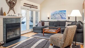 Un salon avec une cheminée, un canapé, un fauteuil à motifs et un tableau représentant un paysage de montagne.