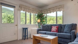 A living room with a sofa, a coffee table, a lamp, and a view of greenery outside. - Sunday River Condo: Brookside I B-306 (Newry)