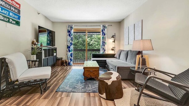 Una sala de estar con sofá, sillón, reposapiés y una mesita de noche con lámpara. Hay un televisor sobre un mueble bajo, una ventana con cortinas y vistas a la vegetación exterior.