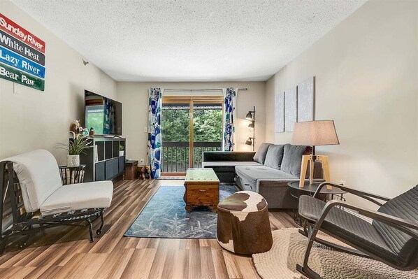 Una sala de estar con sofá, sillón, reposapiés y una mesita de noche con lámpara. Hay un televisor sobre un mueble bajo, una ventana con cortinas y vistas a la vegetación exterior.