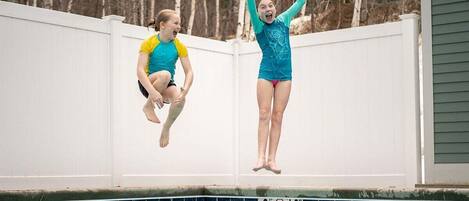 Two individuals jumping into a swimming pool with a white fence and trees in the background.