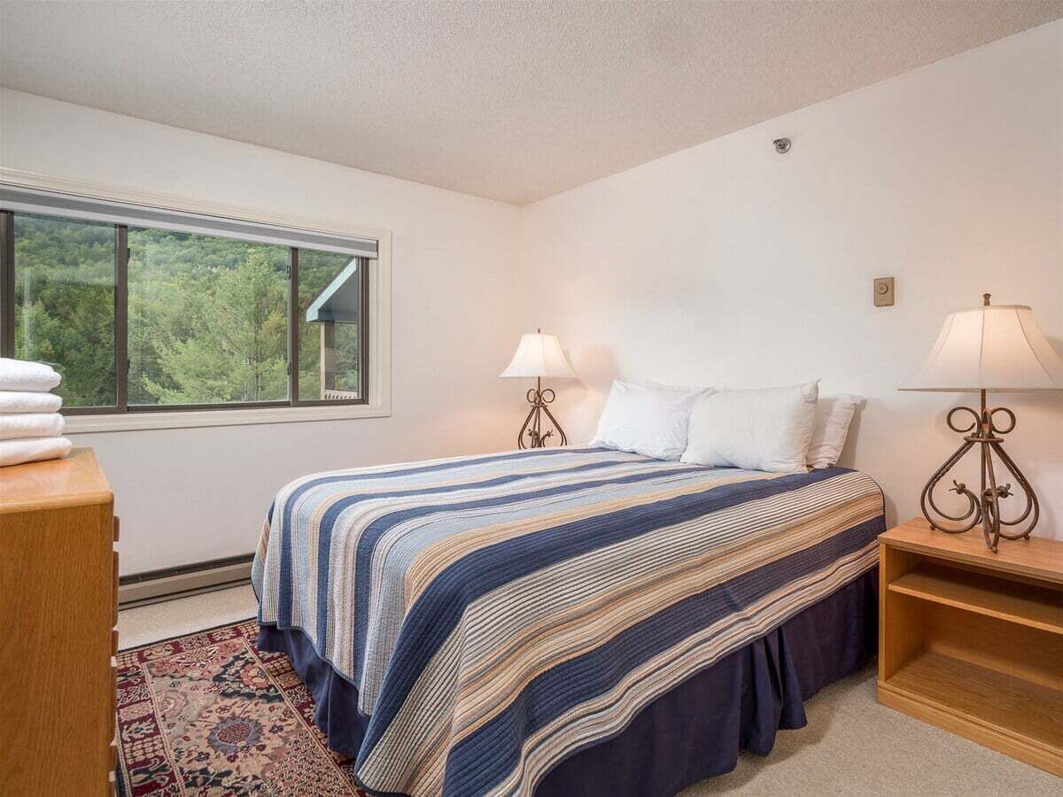 A bedroom with a large window, a bed with striped bedding, two bedside lamps, and a wooden nightstand.
