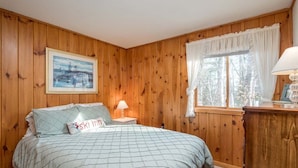 Quarto com painéis de madeira, uma cama, mesa de cabeceira, janela com cortinas e um quadro na parede.