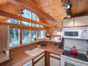 A cabin-style kitchen with wooden beams, a stove, microwave, and a large window overlooking a snowy landscape.
