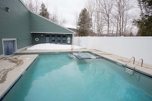 A rectangular swimming pool with a tiled edge, a metal ladder, and a snow-covered area adjacent to a building with a green exterior.
