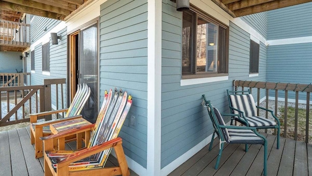 A covered deck with wooden chairs and skis, a wooden railing, and a building with blue siding. - White Cap A-104 - Sunday River Condo (Newry)