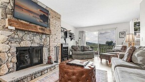 Un salon avec une cheminée en pierre, une grande fenêtre offrant une belle vue, un canapé gris et un pouf rond.