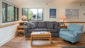 Ein Wohnzimmer mit grauem Sofa, einem blauen Musterstuhl, einem Holztisch und zwei Nachttischen mit Lampen.