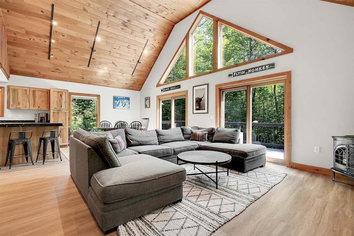 A spacious living area with a large L-shaped sectional sofa, a round coffee table, and a high ceiling with wooden beams. There are large windows providing natural light and views of the outdoors.