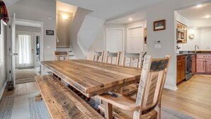 A dining area with a wooden table and chairs, a kitchen in the background, and a door leading to another room.