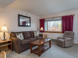 A furnished living room with a sofa, armchair, coffee table, lamp, and a window with curtains. - Sunrise A-131 - Sunday River Condo (Newry)