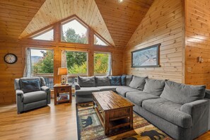 A cozy living room with a sectional sofa, a coffee table, and a fireplace. Large windows offer a view of the outdoors.