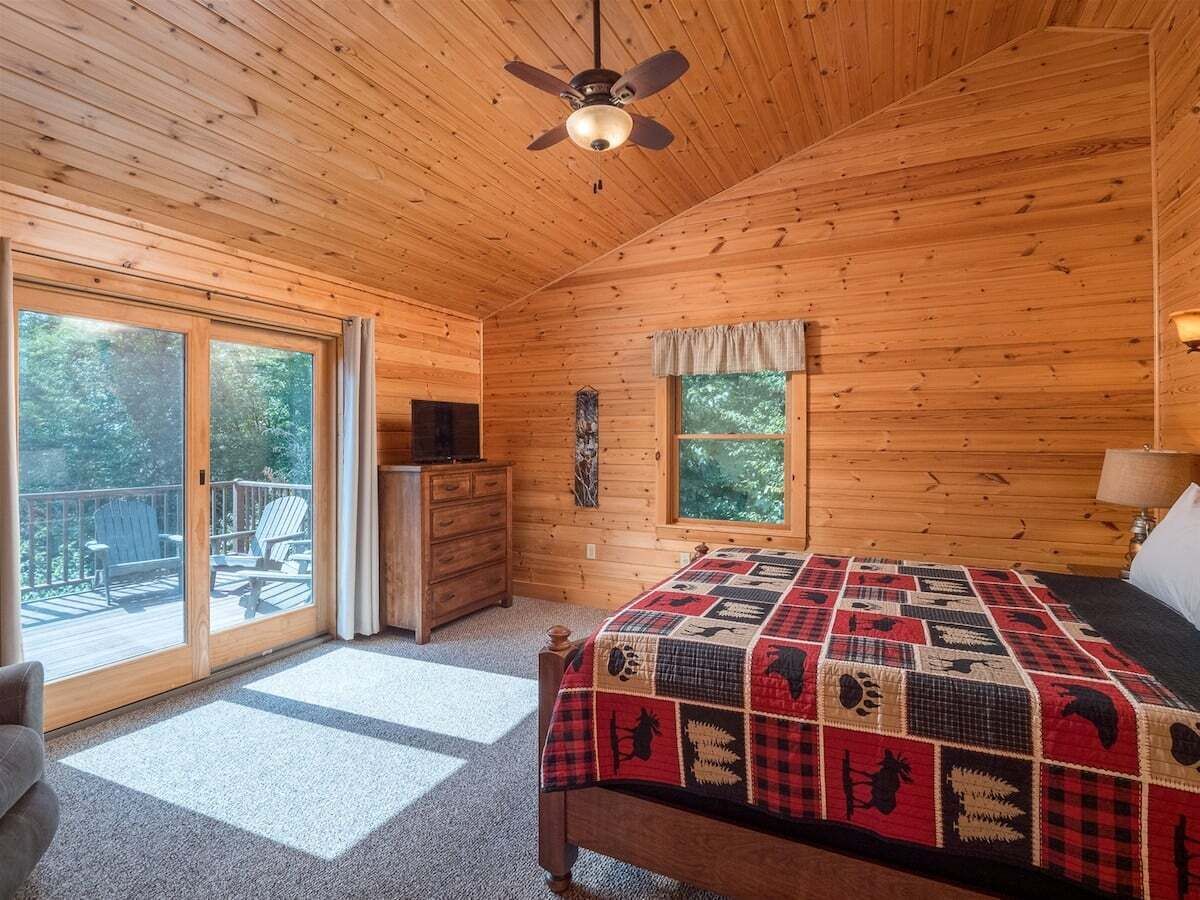 Une chambre dans une cabane en bois, avec un lit, une commode, un ventilateur de plafond et une porte-fenêtre coulissante donnant sur une terrasse extérieure.