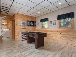 Une pièce aux murs en bois, avec un baby-foot, des cibles de fléchettes et une fenêtre avec des rideaux.
