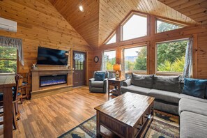 A cozy cabin interior with a fireplace, a television, a sofa, and a dining area.