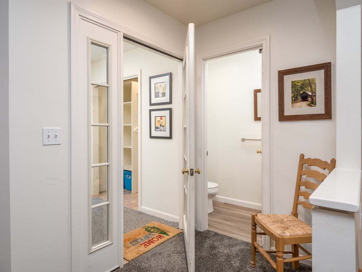 A hallway leading to a bathroom with a toilet, framed pictures on the wall, and a wooden chair.