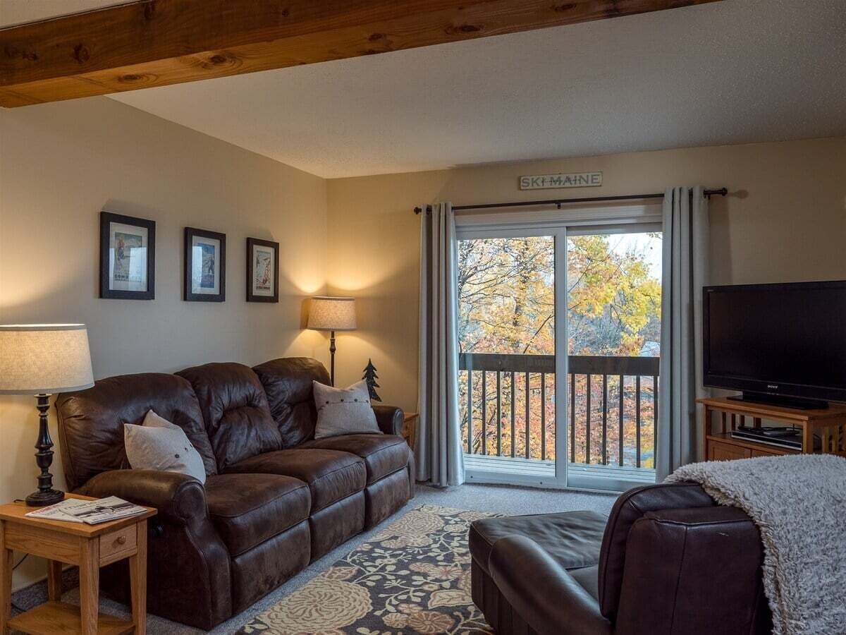 A cozy living room with a leather sofa, a TV, and a large window with a view of trees.