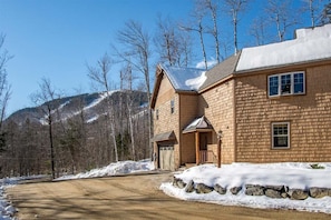 A two-story wooden house with a stone foundation, a garage, and a driveway leading to a snow-covered road. - Timberline Lodge at Sunday River (Newry)